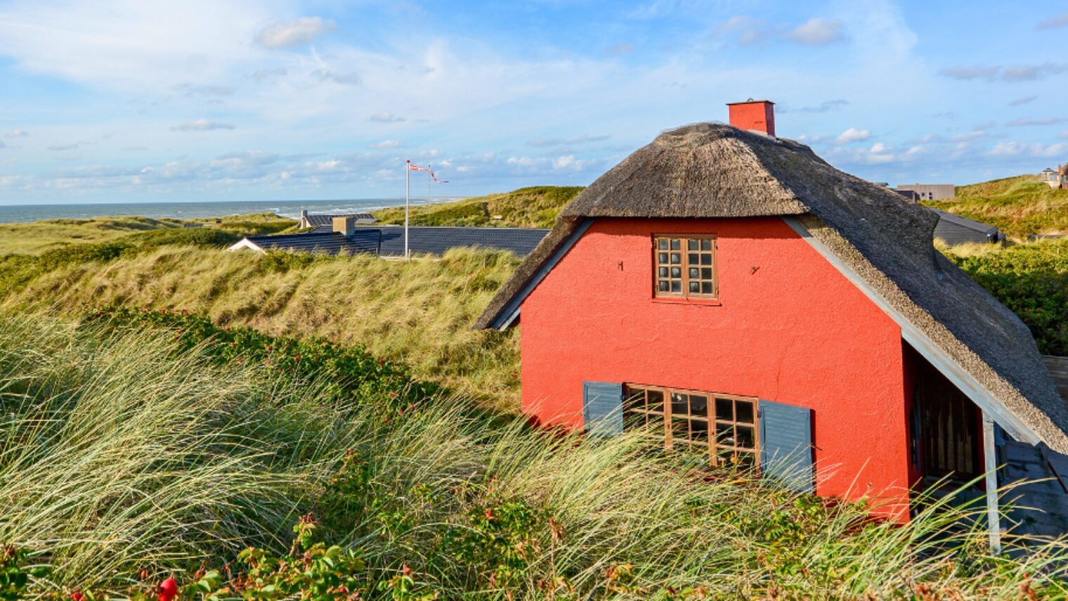 Rotes Haus mit Reetdach in einer Dünenlandschaft. Im Hintergrund ist blauer Himmel zu sehen.