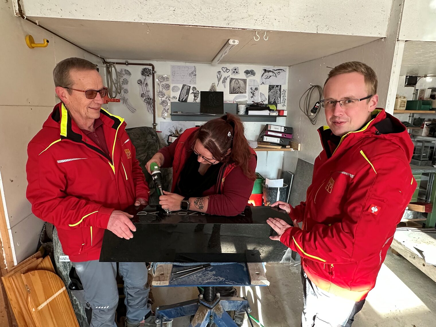 Zwei Handwerker und eine Handwerkerin stehen in der Werkstatt und bearbeiten einen Grabstein