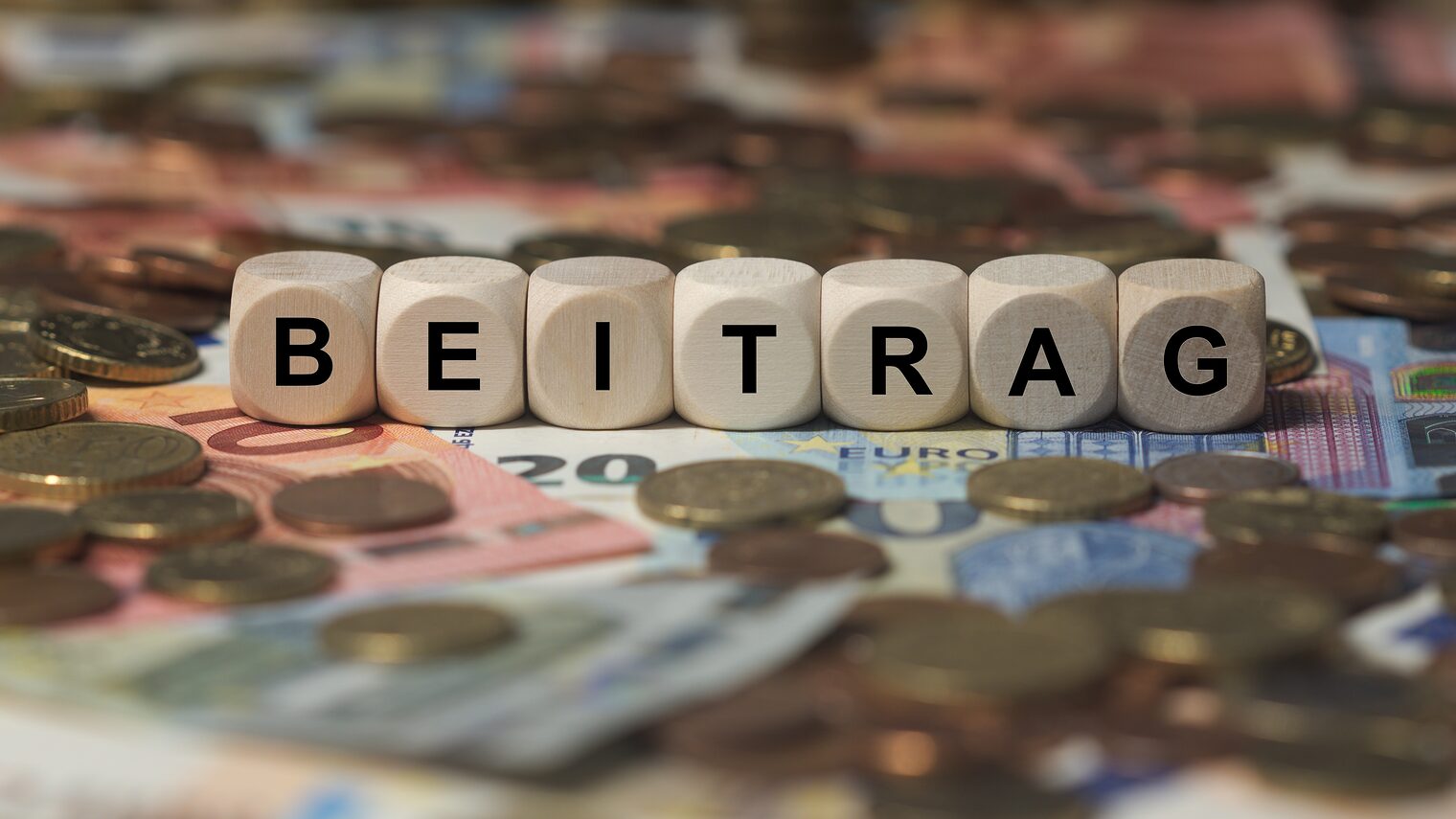 Holzwürfel mit dem Wort „Beitrag“ auf Euro-Banknoten und Münzen als Symbol für Beiträge und Finanzierung.